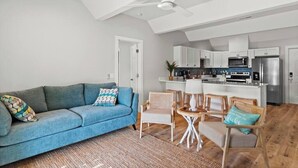A modern living room with a blue sofa, a dining area with wooden chairs, and a kitchen with white cabinets and stainless steel appliances.