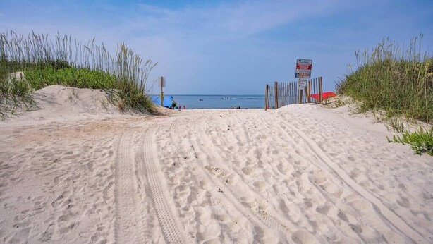 A sandy beach path leading to the sea with a signpost and a red umbrella on the right.