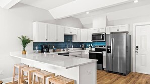 A modern kitchen with white cabinets, a large island with stools, a stainless steel refrigerator, and a microwave.