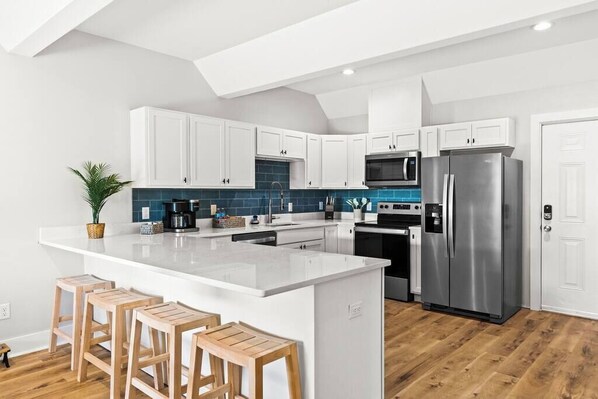 A modern kitchen with white cabinets, a large island with stools, a stainless steel refrigerator, and a microwave.