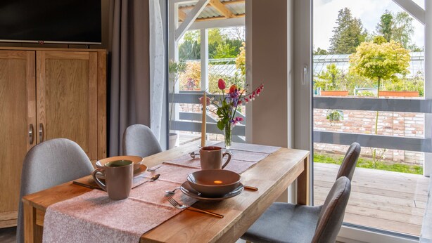 Een eetgedeelte met een houten tafel, stoelen, een vaas met bloemen en een televisie aan de muur.
