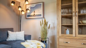 Een moderne woonkamer met een grijze bank, een glazen salontafel, een houten kast met glazen deuren, een vloerlamp en een ingelijste foto aan de muur.