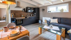 Een moderne keuken met donkere kasten, een gasfornuis en een koffiezetapparaat. Er is een eethoek met een glazen tafel en stoelen, en een woonkamer met een grijze bank en een raam met uitzicht op groen.