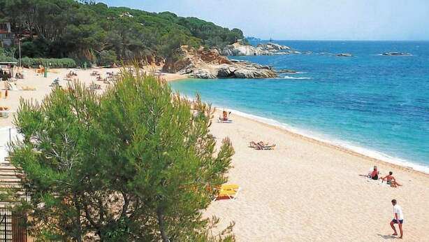 Une plage avec une eau d’un bleu limpide, un rivage sablonneux et des affleurements rocheux.