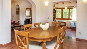 Un coin repas avec une table ronde en bois et des chaises, une sculpture de coq blanche, et une vue sur la cuisine et le salon, visibles à travers une arche.