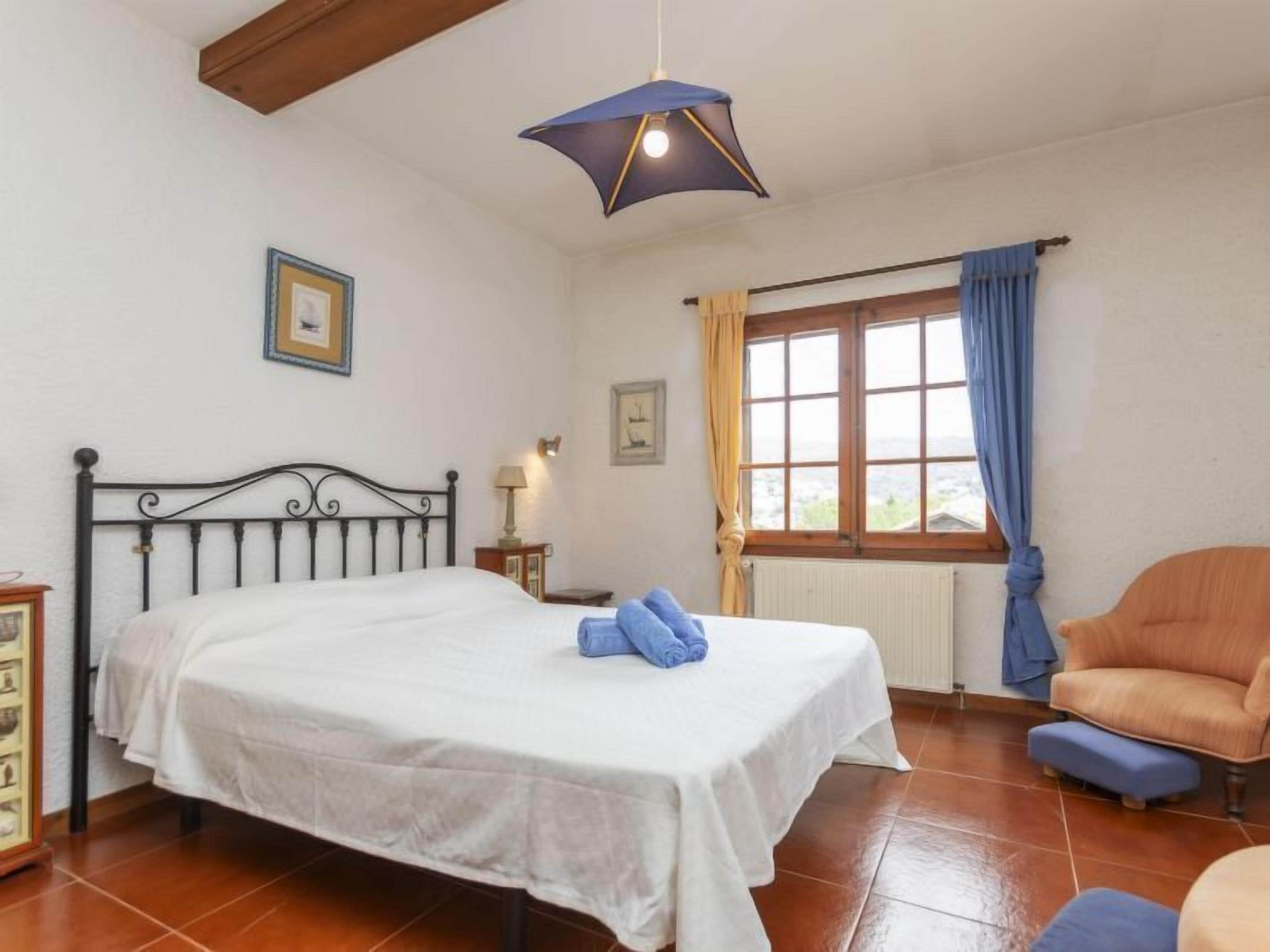 Ein Schlafzimmer mit Bett, Sessel, einem Fenster mit Vorhängen und einer Pendelleuchte.