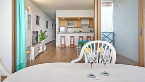Ein moderner Essbereich mit einem weißen Tisch und Stühlen, eine Küche im Hintergrund und Meerblick durch eine Schiebetür.