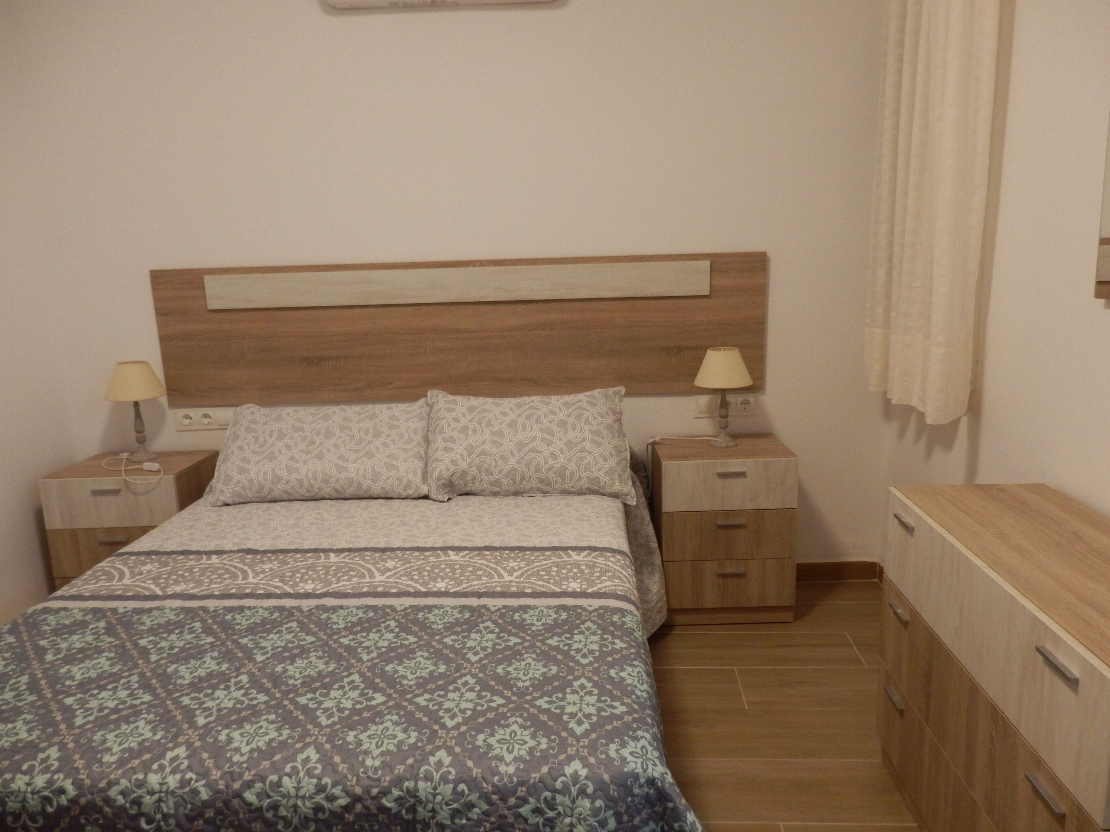 A bedroom with a wooden headboard, two bedside tables with lamps, a bed with patterned bedding, and a wooden dresser.