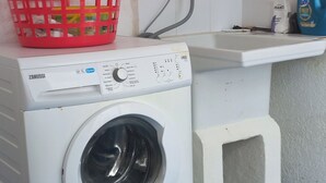 Un espace buanderie compact comprenant un lave-linge blanc, un panier à linge rouge et un petit évier avec un tuyau.