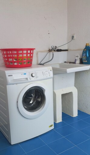 Espaço compacto de lavanderia com máquina de lavar branca, cesto de roupa vermelha e uma pia pequena com uma mangueira.