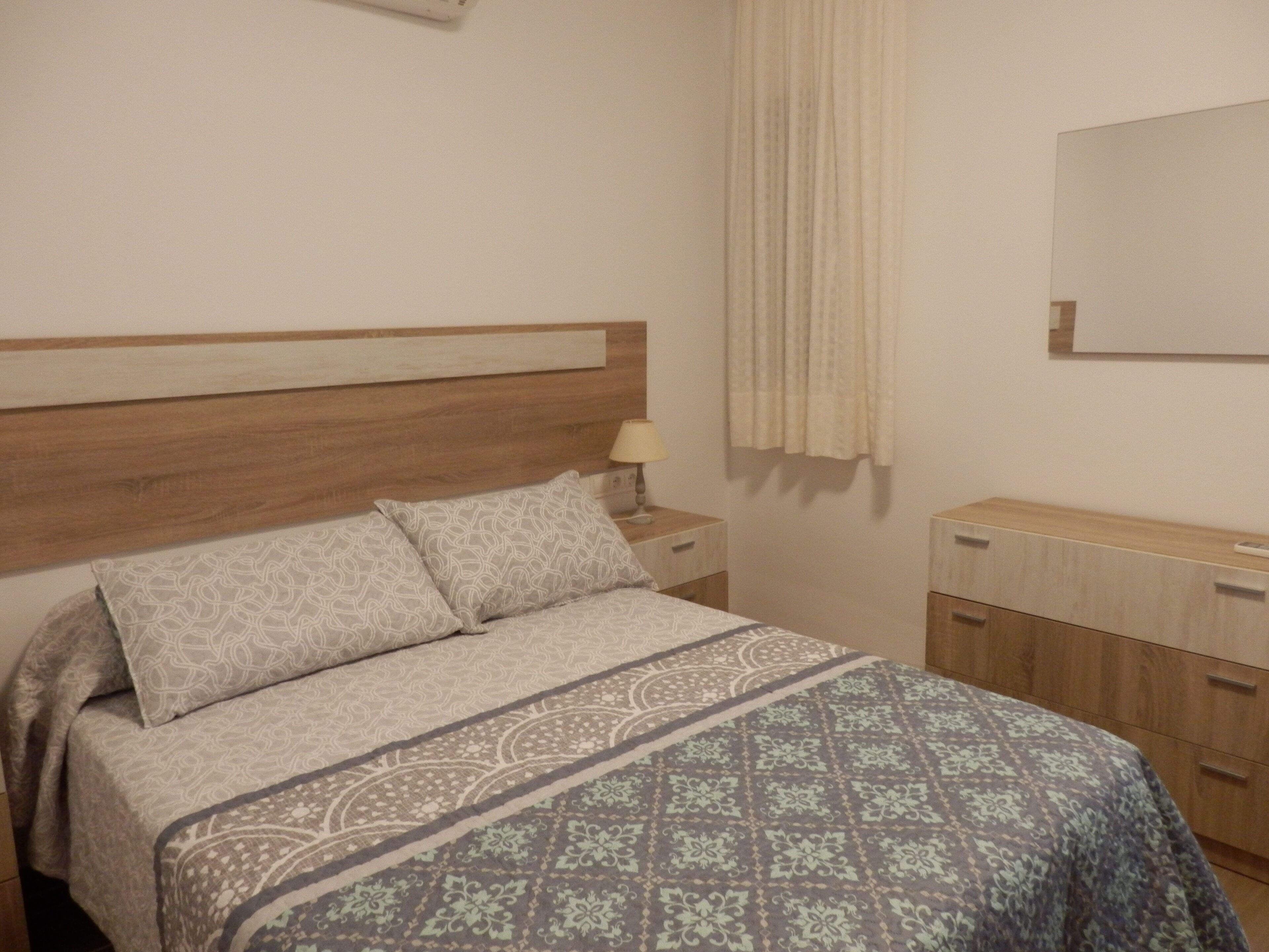 Quarto com cabeceira de madeira, cama com roupa de cama estampada, mesa de cabeceira com abajur, espelho e cômoda de madeira.