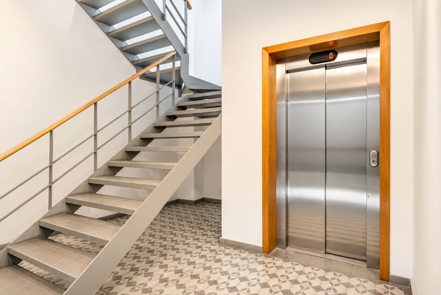 A modern elevator with a digital display and a wooden frame, adjacent to a staircase with a metal railing and patterned floor tiles.