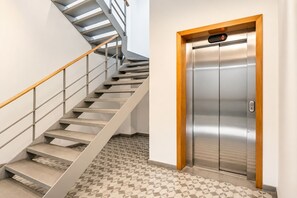 A modern elevator with a digital display and a wooden frame, adjacent to a staircase with a metal railing and patterned floor tiles.