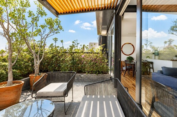 A modern outdoor patio with wicker furniture, a glass door leading to a dining area, and a view of the sky and trees.