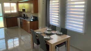 Une cuisine moderne avec des meubles en bois, une table à manger pour quatre personnes et une grande fenêtre équipée de stores.