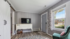 Una habitación con puerta corredera, un tocador blanco con televisor, un sillón verde y una alfombra estampada.