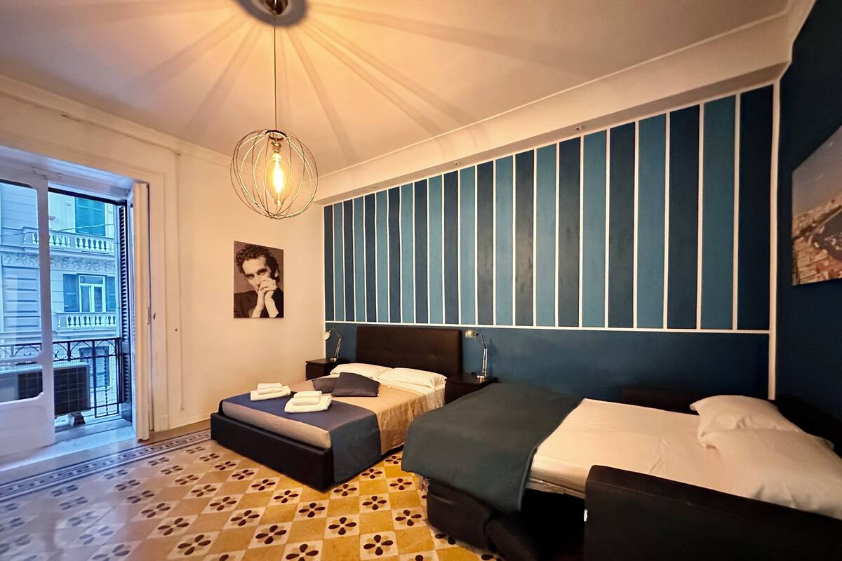 A modern bedroom with a large bed, a smaller bed, a nightstand, a lamp, and a view of the city through a balcony.
