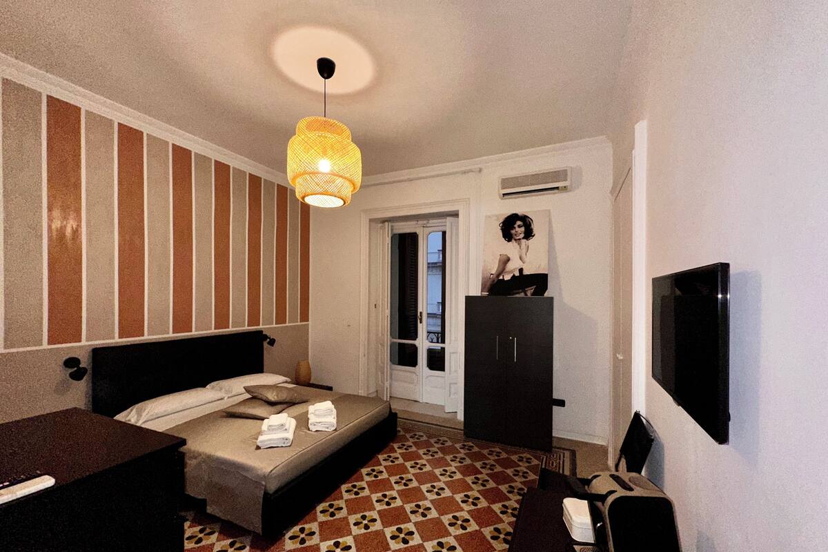 A hotel room with a bed, a desk, a wardrobe, a television, and a patterned floor.