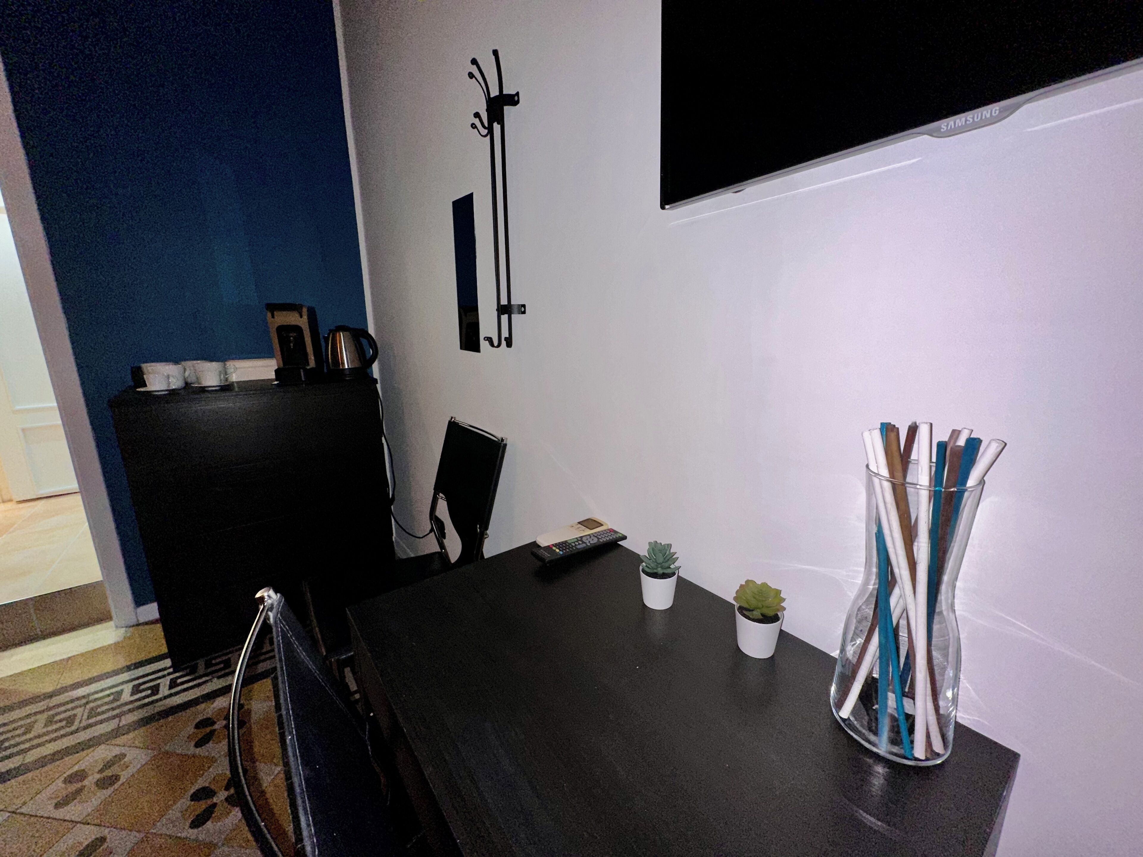 Un coin repas avec une table en bois foncé, deux petites plantes en pot, un vase en verre contenant des stylos, une chaise noire et un meuble noir abritant une bouilloire et des tasses.