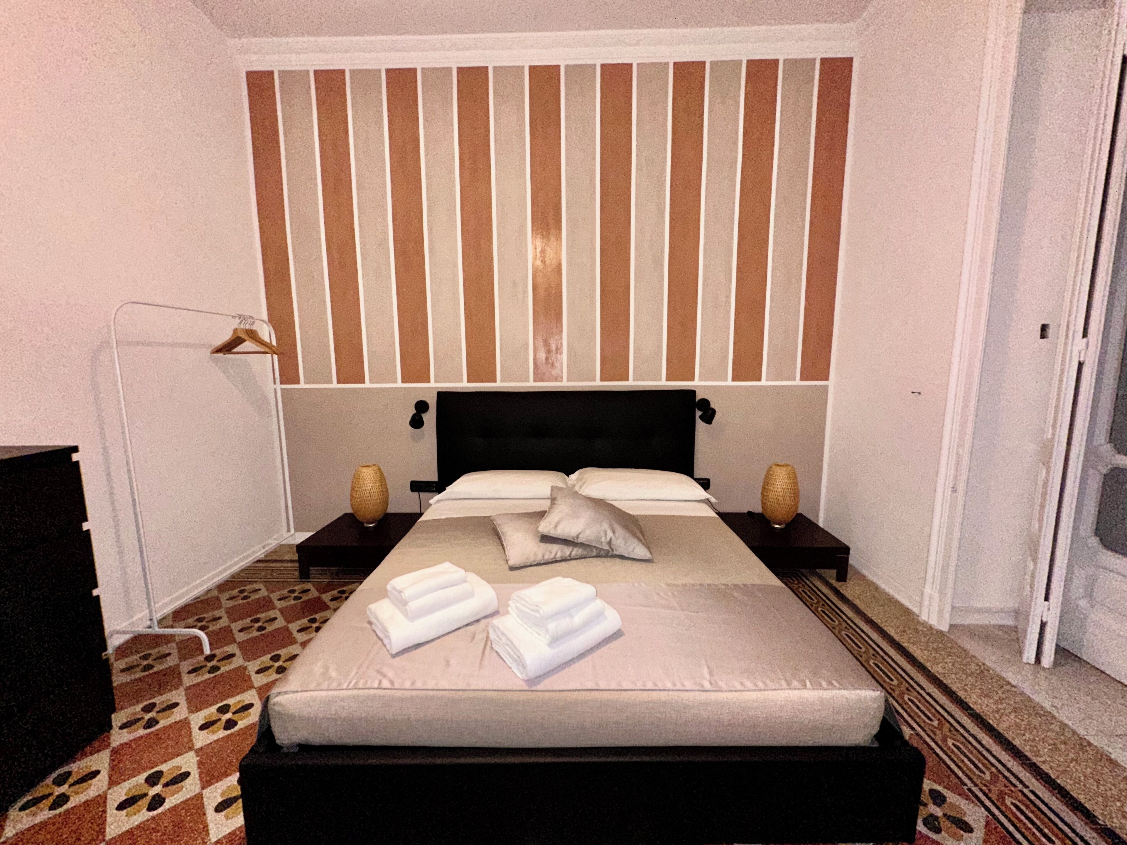 A neatly made bed with white linens and pillows, a bedside table with a lamp and a vase, and a patterned carpet.