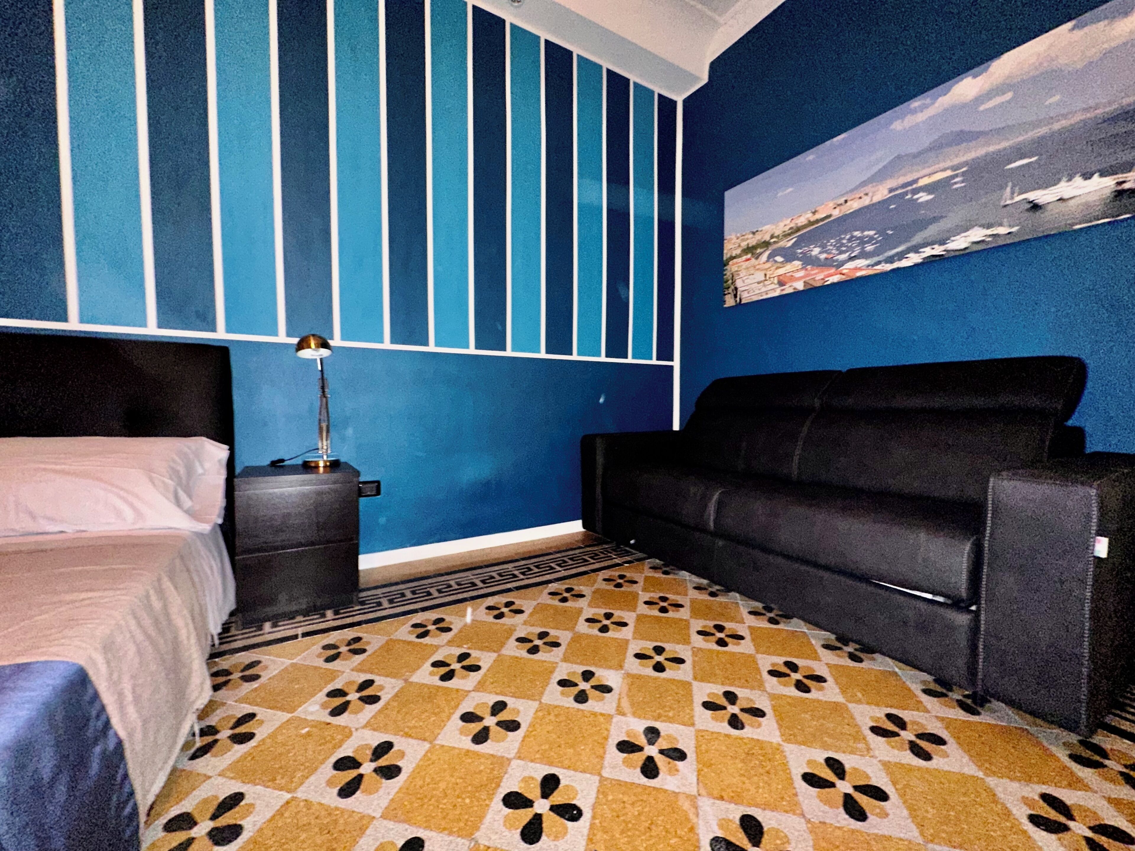 A room with a bed, a black sofa, a desk with a lamp, a patterned floor, and a painting of a coastal scene.
