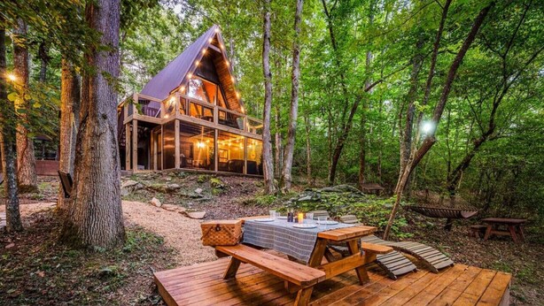 A cabin with a sloped roof, surrounded by trees, featuring a deck with benches and a table set for dining. - NEW! Creekside Ranch A-Frame (Young Harris)