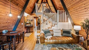 A cozy loft-style room with a wooden interior, a brown leather sofa, a dining table, and a staircase leading to an upper level. - NEW! Creekside Ranch A-Frame (Young Harris)