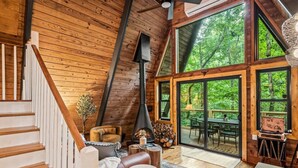 A cozy cabin interior with a wooden interior, a fireplace, and large windows offering a view of the surrounding forest. - NEW! Creekside Ranch A-Frame (Young Harris)