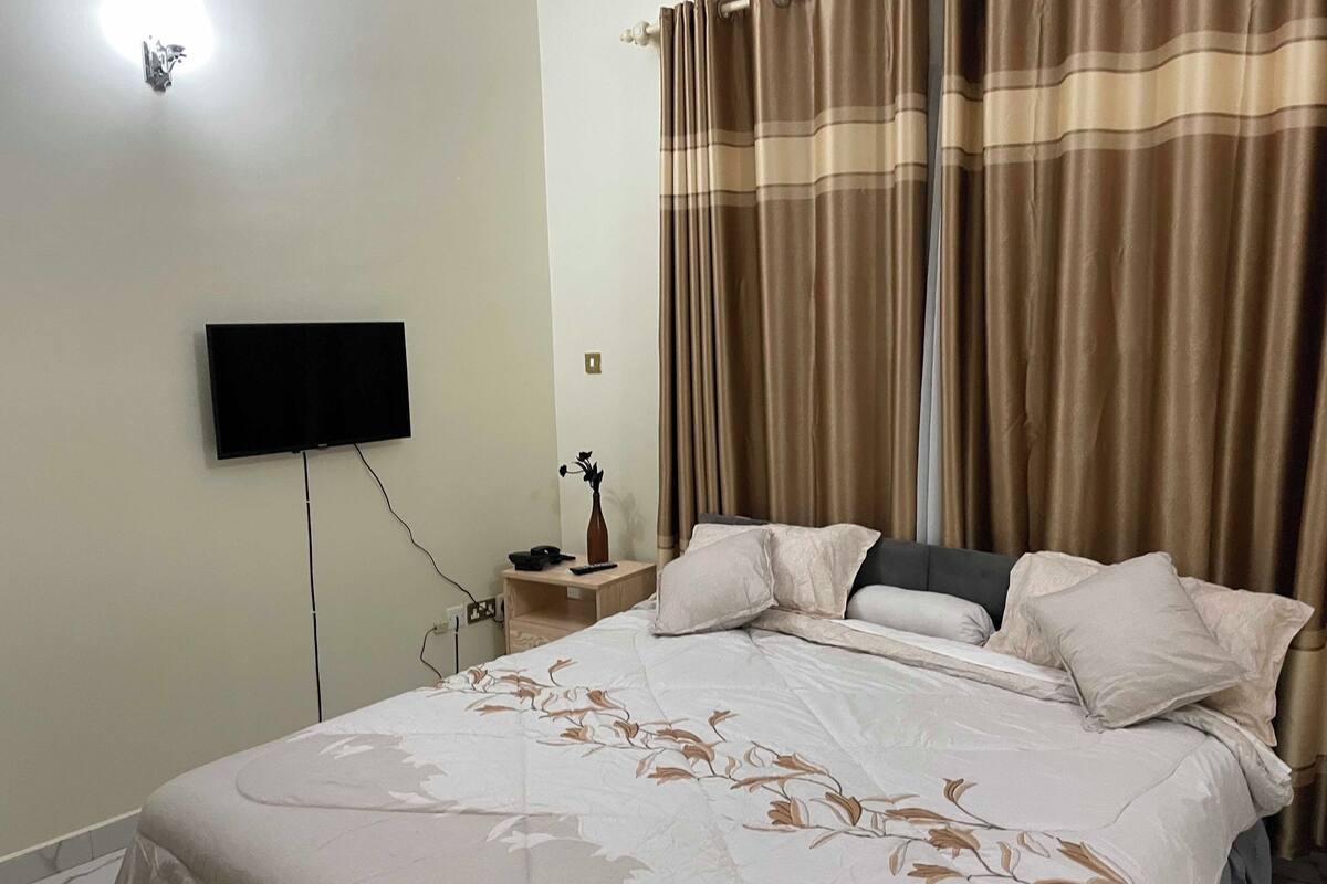 Ein Schlafzimmer mit Bett, einem an der Wand befestigten Fernseher, einem Nachttisch mit Vase und Vorhängen.