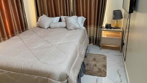 A neatly made bed with a beige comforter and pillows, a bedside table with a lamp, and a TV mounted on the wall. - Kampala (Kampala)