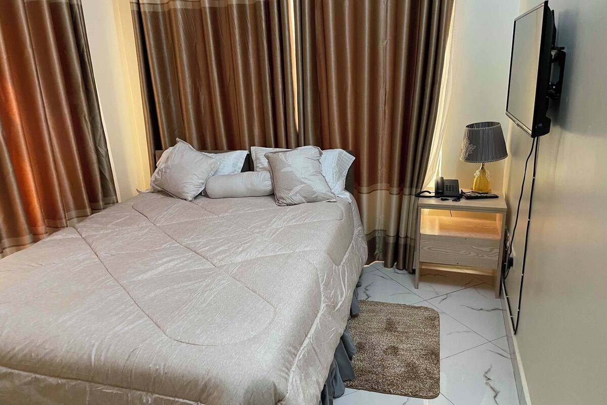 Ein ordentlich bezogenes Bett mit einer beigen Steppdecke und Kissen, ein Nachttisch mit Lampe sowie ein an der Wand befestigtes Fernsehgerät.