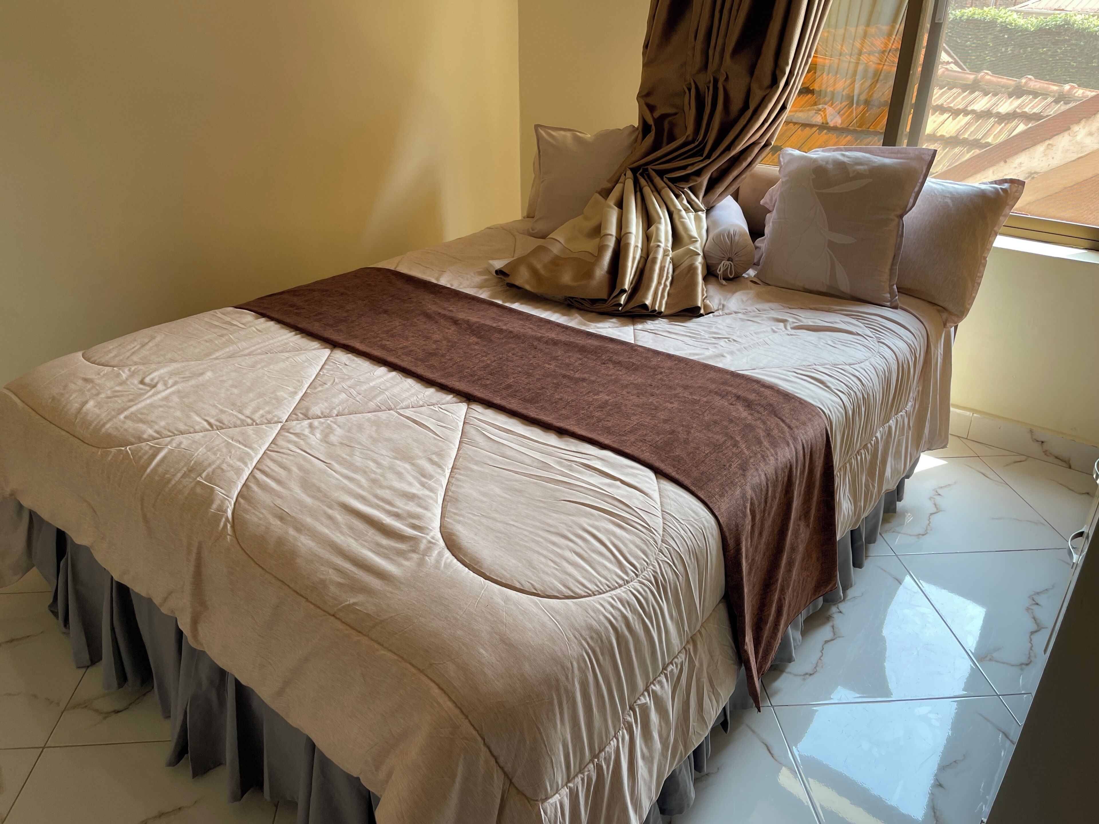 A neatly made bed with a beige quilt and a brown throw blanket, a window with curtains, and a tiled floor.