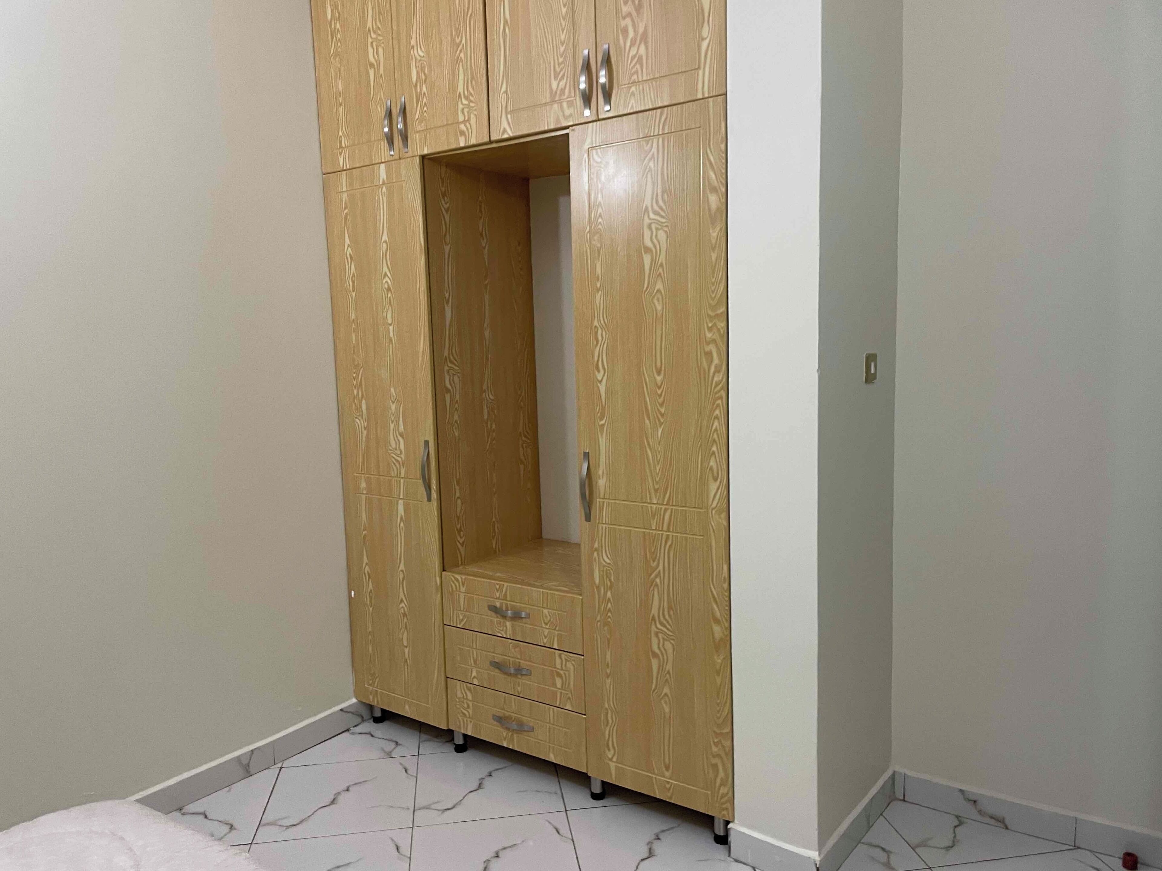Una habitación con un armario de madera con varios cajones y puertas, suelo de baldosas con diseño de mármol y paredes lisas.