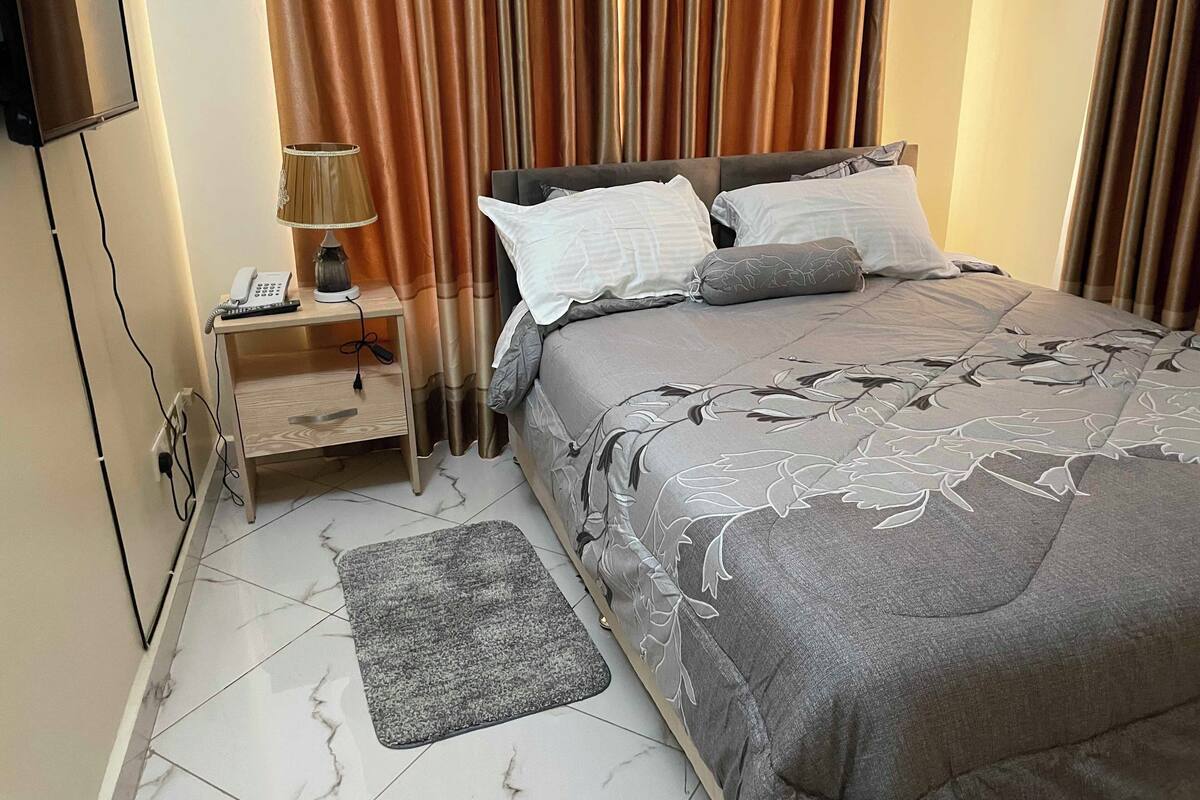 Ein ordentlich eingerichtetes Schlafzimmer mit einem Bett, Kissen, einem Nachttisch mit Lampe und einem Telefon.