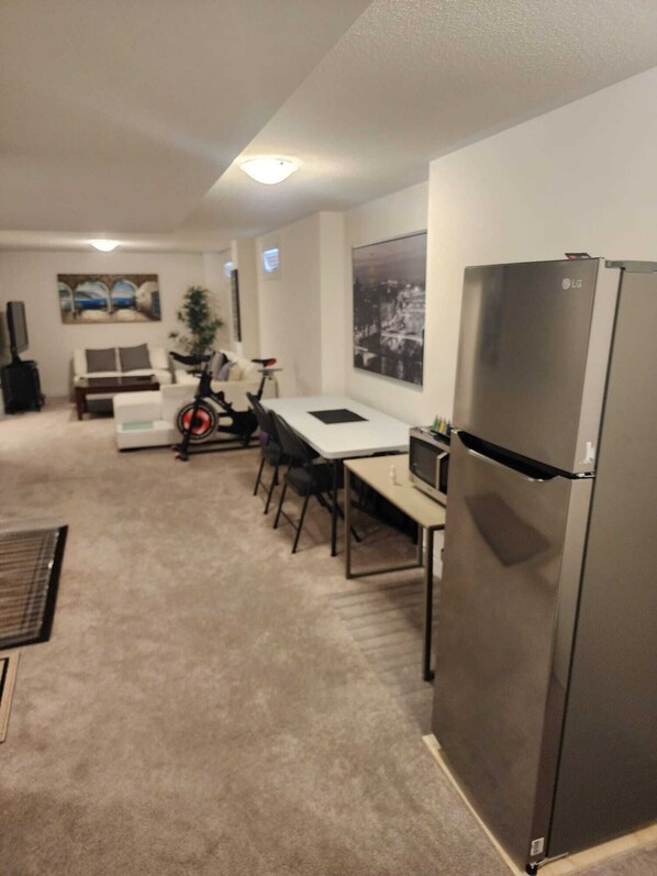 A compact living space with a kitchenette, a dining area with a table and chairs, a small sofa, a bicycle, and a refrigerator. - Cozy Unit (2 Rooms), King & Double Bed Perfect for Couples, Singles & Families (Brampton)