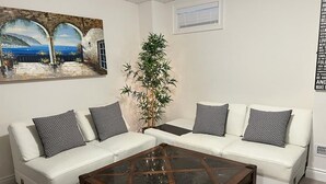 Un salón con un sofá blanco, una mesa de centro de cristal y un cuadro de una escena costera.