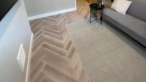 Un salon moderne avec un parquet en chevrons, un canapé gris, un tapis à motifs et un téléviseur fixé au mur.