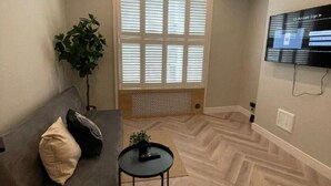 Un salon moderne avec un canapé gris, une petite table ronde, une plante en pot et une grande fenêtre avec des stores.
