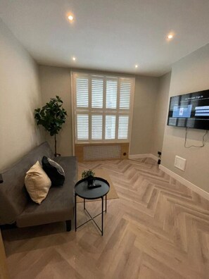 Relax in this stylish living room with a sofa, side table, and potted plant, surrounded by a herringbone-patterned floor and a flat-screen TV.