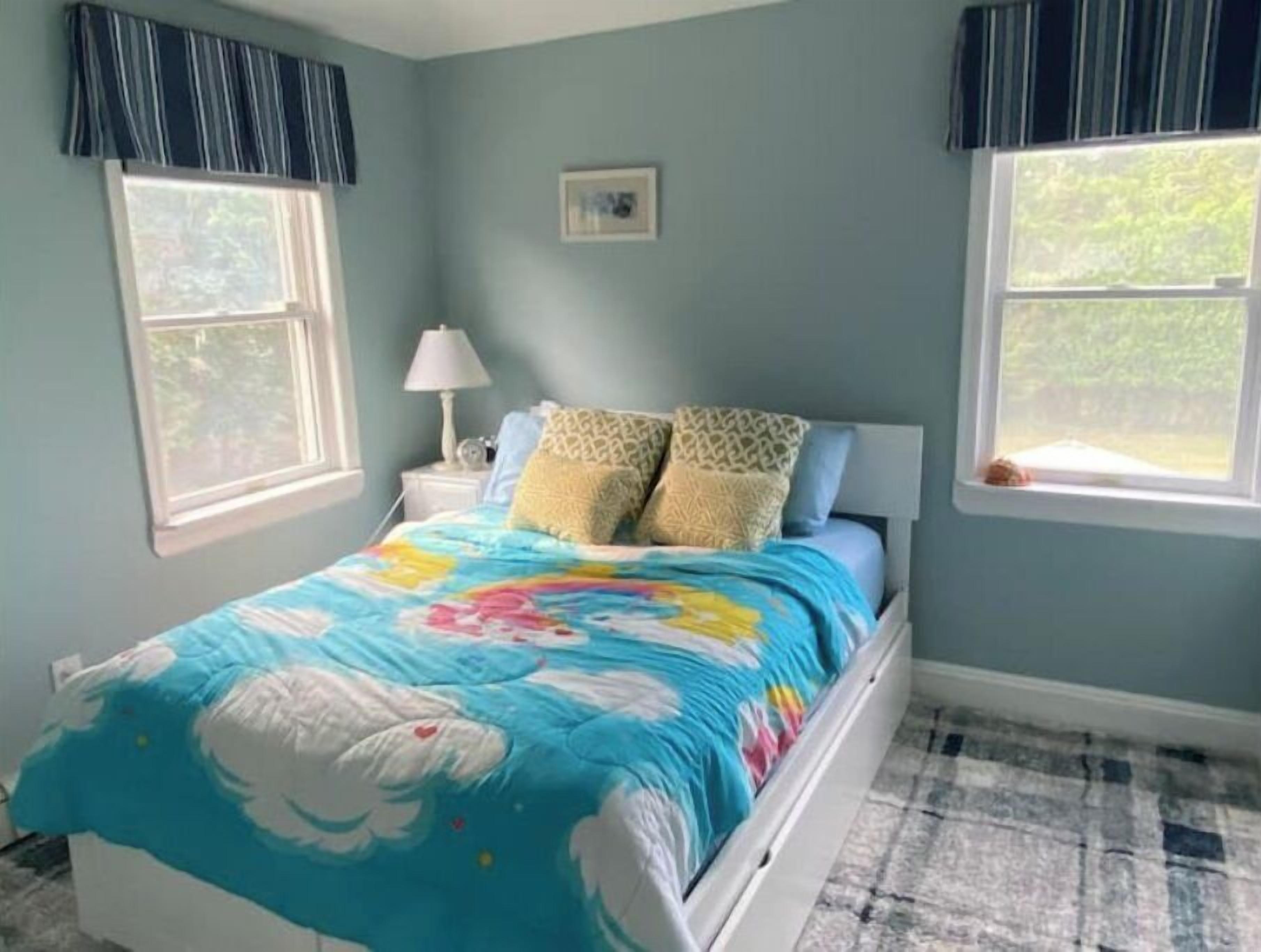 A bedroom with a bed, two windows, a nightstand, and a lamp.