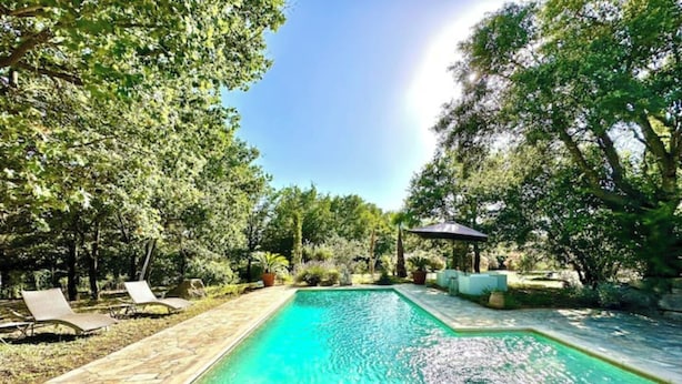 A rectangular swimming pool with a stone border, surrounded by trees and a patio area with lounge chairs. - Villa Julien (Gassin)