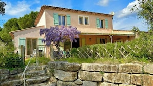 A two-story house with a stone wall in the foreground, a wooden fence, and a purple flowering tree in the front yard. - Villa Julien (Gassin)