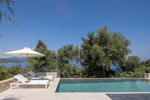 A swimming pool with lounge chairs and an umbrella, surrounded by trees and a view of the sea.
