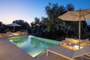 Piscina con sillas de descanso y sombrillas, rodeada de árboles.