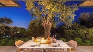 Un espacio de comedor al aire libre con una mesa preparada para dos, rodeado de sillas de mimbre, bajo una pérgola de madera con un árbol en el centro.