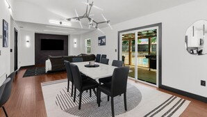 A modern living room with a dining area, a large TV, and a sliding glass door to a backyard. - Adventure Coast Modern; 3/3 Pool w/ Firepit, Fenced, Heated Pool (Spring Hill)