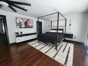 A modern bedroom with a four-poster bed, a large painting, a white dresser, and a dark wooden floor.
