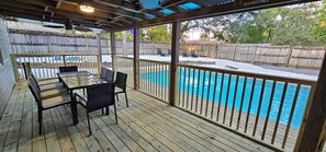 A covered outdoor area with a pool, wooden deck, and a table with chairs. - Adventure Coast Modern; 3/3 Pool w/ Firepit, Fenced, Heated Pool (Spring Hill)