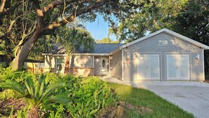 A two-story house with a garage, surrounded by lush greenery and large trees. - Adventure Coast Modern; 3/3 Pool w/ Firepit, Fenced, Heated Pool (Spring Hill)