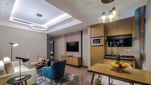 Una sala de estar moderna con un sofá, una mesa de centro, una silla azul y una mesa de comedor con un frutero. Hay un televisor de pantalla plana montado en la pared, un microondas y una lámpara del techo.